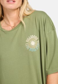 Ylihuliseva oliivivihreä t-paita pehmeästä puuvillasta, jossa on pyöreä kaula-aukko ja edessä graafinen printti auringosta, jossa on teksti "Billabong World Tour 1973".