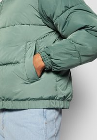 Chaqueta acolchada en verde apagado, con un cuello alto, puños elásticos, bolsillos laterales y un diseño acolchado con una textura suave.