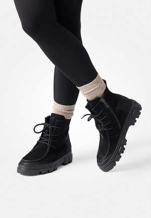 Ben i sorte leggings iført beige sokker og sorte snørestøvler med chunky såler, stående på hvid baggrund.