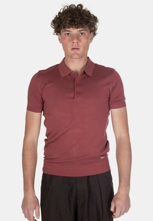 Giovane uomo con capelli ricci che indossa una polo aderente a maniche corte di colore rosso mattone e pantaloni scuri, in piedi davanti a uno sfondo uniforme.