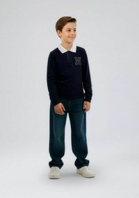 Polo de manga larga azul marino con cuello blanco y una letra "N" bordada en azul. Combinado con vaqueros de corte holgado azules y zapatillas blancas.