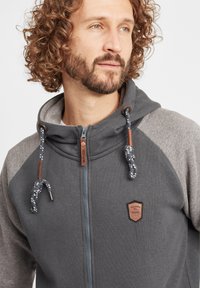 Men's hooded sweatshirt in dark gray with light gray raglan sleeves. Features a zip closure, decorative logo, and rope drawstrings.