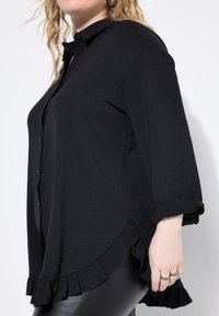 Camisa negra de botones con una tela texturizada, mangas enrolladas y un dobladillo con volantes, que proporciona una silueta relajada y casual.