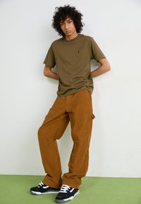 Pantaloni din corduroy maron, tricou scurt cu mânecă scurtă de culoare verde măslină și adidași negri cu șireturi albe. Croi slim, material texturat, design simplu.