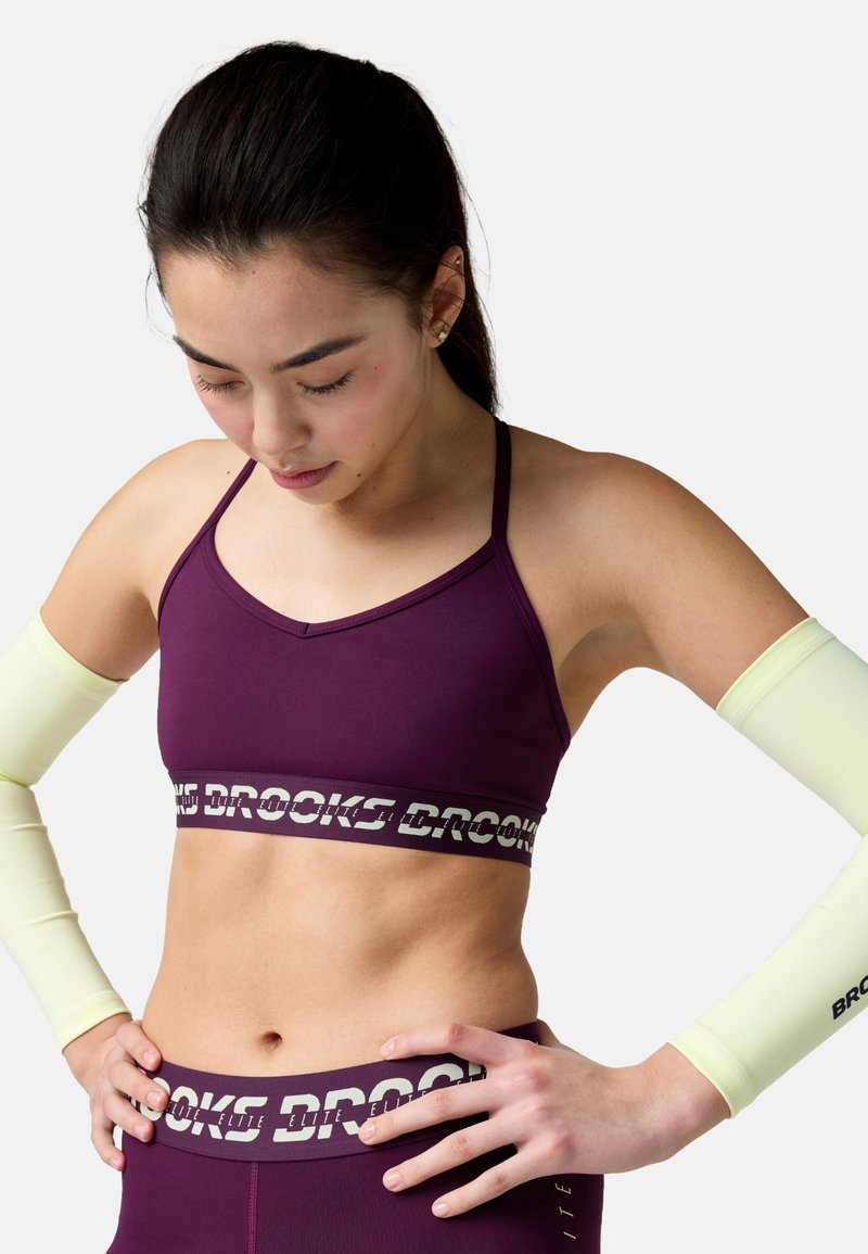 Naine burgundiaspordirinnahoidja ja retuusidega, millel on "Brooks" logo, kandes helekollaseid käevarsi, vaatab alla, käed puusal, valge tausta ees.