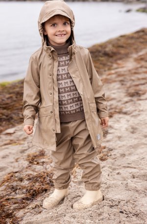 Veste imperméable beige avec capuche, assortie à un pantalon et des bottes en caoutchouc. Le pull présente un motif "M" marron répétitif.