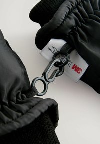Schwarze isolierte Handschuhe mit einem strukturierten Stoffbereich am Handgelenk und einem schwarzen Befestigungsclip. Enthält ein sichtbares 3M-Etikett, das in das Material eingenäht ist.