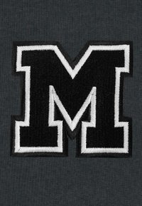 Crno izvezeno slovo "M" obrubljeno bijelom bojom na tamnoj teksturiranoj pozadini. Dizajn se ističe odvažnim linijama i strukturiranim oblikom.