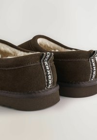 Pruunid nahast slip-on kingad, millel on pehme sisu, detailne õmblus ja tekstureeritud punutud aktsent kontsal. Lame talla disain.