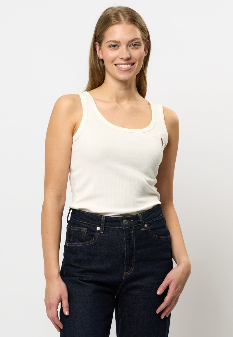 Vit ribbad linne med breda axelremmar och rund halsringning, med en liten broderad detalj på bröstet, kombinerad med mörkblå jeans.