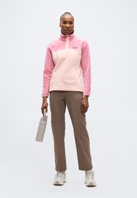 Γυναίκα που στέκεται φορώντας ροζ φλις πουλόβερ, καφέ παντελόνι, λευκά αθλητικά παπούτσια, κρατώντας ένα γκρι παγούρι νερού σε απλό ανοιχτό φόντο.