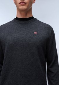 Pull gris foncé en tissu doux, avec un col rond et un petit emblème du drapeau norvégien sur le côté gauche de la poitrine.