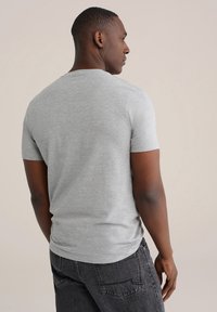 Mees vasakule poole vaadates, kandes lihtsat helehallist lühikese varrukaga t-särki ja musta teksapükse beeži tausta ees.