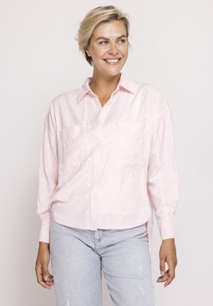 Smiling woman with short blonde hair wearing a loose pink striped button-up shirt and light blue jeans, standing against a plain background.