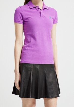 Kvinde iført en tætsiddende lilla poloshirt og et sort plisseret lædernederdel, står mod en ensfarvet lys baggrund. Ansigtet er ikke synligt.