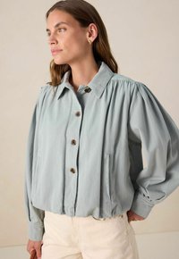 Veste boutonnée bleu clair avec des manches bouffantes, deux poches latérales et des boutons en bois. Tissu doux et texturé avec un ourlet court.