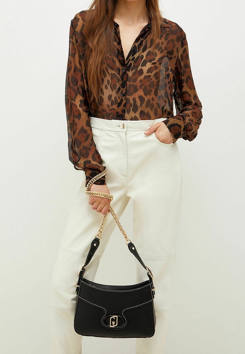 Femme portant un chemisier marron transparent à imprimé léopard et un pantalon blanc, tenant un sac à main noir avec une bandoulière en chaîne dorée.