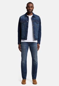 Giacca di jeans blu scuro con chiusura a bottoni, due tasche sul petto e cuciture a contrasto, indossata sopra una maglietta bianca e jeans blu.