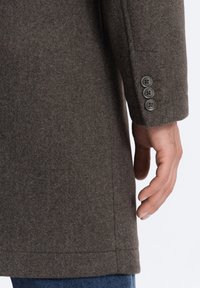 Manteau en laine gris au design ajusté, avec une texture lisse, une coupe droite et des détails de manche à trois boutons. Main reposant sur le bord du manteau.