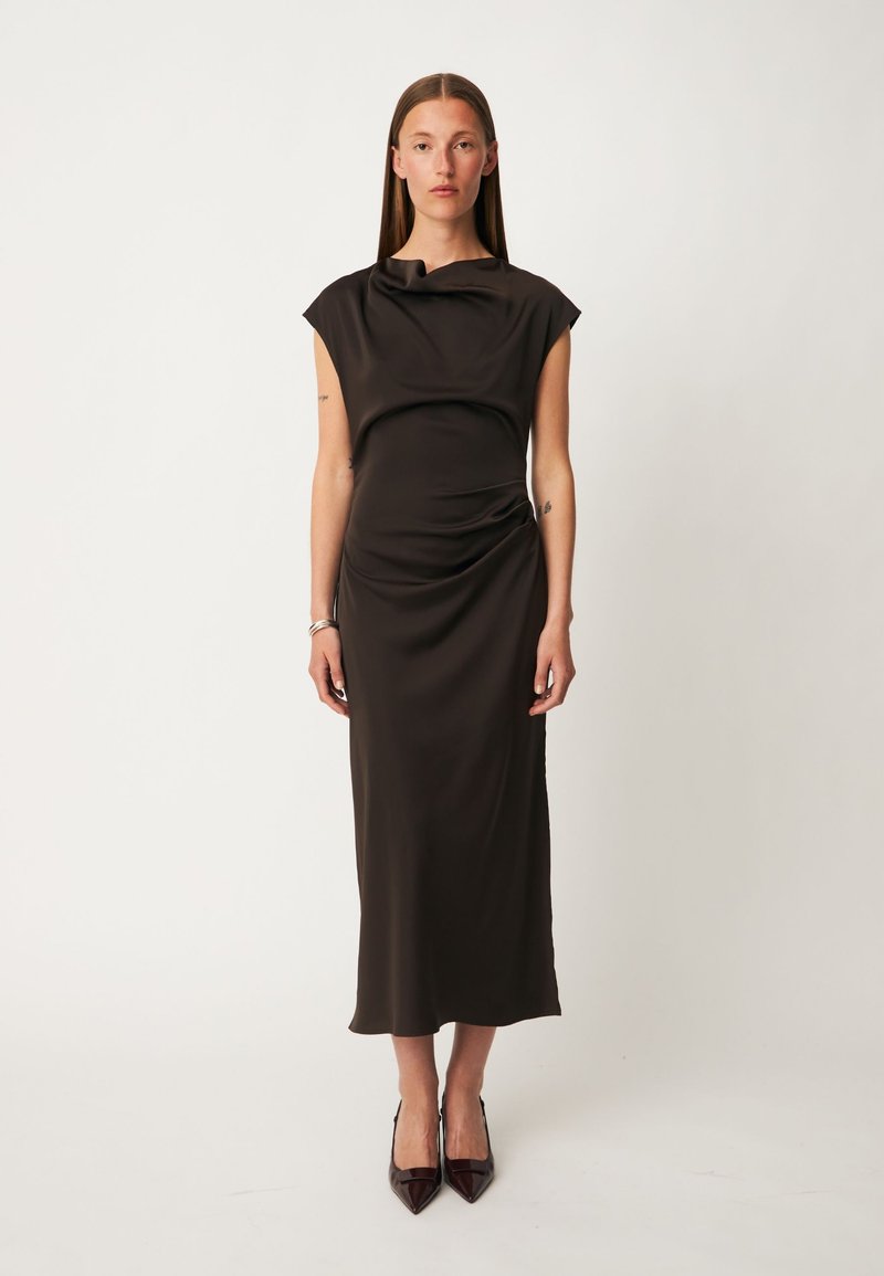 Femme debout portant une robe midi noire sans manches avec des détails drapés et des escarpins noirs pointus sur un fond clair uni.