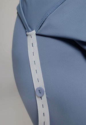 Close-up van een blauwe stoffen kledingstuk met een witte elastische band met stikdetails en een ronde paarse knop aan de zijkant.