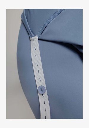 Close-up van een blauwe stoffen kledingstuk met een witte elastische band met stikdetails en een ronde paarse knop aan de zijkant.