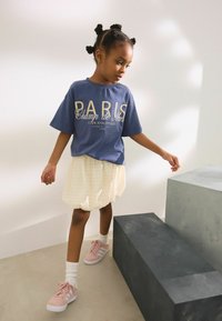 T-shirt bleu avec l'imprimé "PARIS", associé à une jupe crème texturée et des baskets roses avec des rayures blanches. Des chaussettes blanches à côte complètent la tenue.