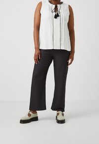 Bílý bezrukávový top s kravatovým dekoltem a černým lemem; spárovaný s černými texturovanými širokými kalhotami a krémovými botami se sponami.