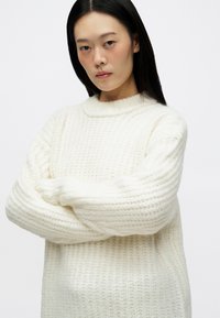 Νεαρή γυναίκα με μακριά σκούρα μαλλιά, φορώντας ένα λευκό χοντρό πλεκτό πουλόβερ, με τα χέρια σταυρωμένα μπροστά σε έναν απλό λευκό φόντο.