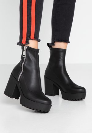 Bottines noires à plateforme avec talons carrés et fermetures éclair latérales, portées avec un pantalon noir à ourlet effiloché arborant des rayures rouges.