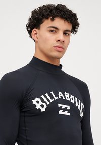Czarny rash guard z wysokim kołnierzem i białym logo "Billabong". Gładki, elastyczny materiał z dopasowanym krojem i płaskimi szwami.