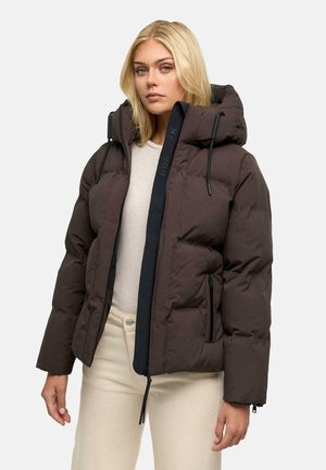 Vrouw draagt een donkerbruine donsjacket met een grote capuchon, beige broek en een lichtbeige top, staand tegen een effen lichte achtergrond.