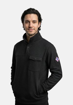 Jonge man met donker haar, gekleed in een zwarte trui met een voorvak en een logopatch op de linker mouw, staand met handen in de zakken.