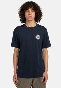 Navy Baumwoll-T-Shirt mit kurzen Ärmeln, Rundhalsausschnitt und Logo-Detail auf der Vorderseite. Glatte Textur und lässige Passform. Kombiniert mit khakifarbenen Hosen.