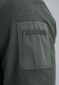 Donkergroene textuurstof met een patchzak. De zak heeft horizontale stiksels en een lus voor het vastzetten van items.