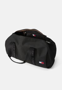 Fekete nejlon sporttáska dupla fogantyúval, cipzárral zárható, egyik oldalán egy kis, színes logófolttal. Belseje barna szövet.