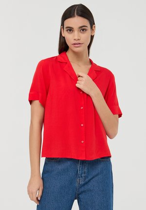 Camicia rossa a maniche corte con colletto, vestibilità comoda e texture morbida, abbinata a jeans blu a vita alta.