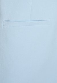 Tejido azul claro con una textura suave, que presenta un bolsillo de canto horizontal integrado en el diseño. Construcción simple y limpia.