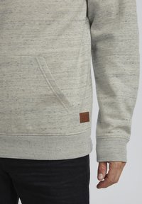 Sweat-shirt gris clair avec un tricot texturé, doté d'une poche kangourou, de poignets côtelés et d'une étiquette en cuir marron au bas.