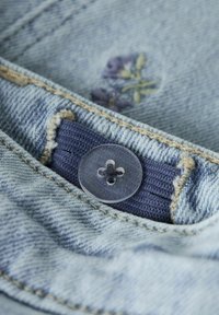 Gai zils džinsis ar bēšām diega šuvēm, kuram ir tumši zilas auduma etiķete un caurspīdīga poga uz kabatas malas.