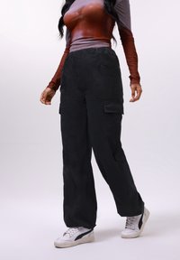 Pantalon cargo noir en tissu lisse, avec poches latérales, coupe décontractée et taille à cordon. Associé à des baskets blanches.