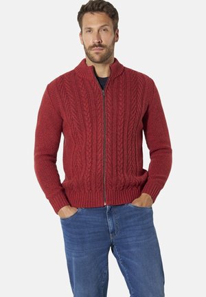 Mann trägt roten Strickpullover mit Reißverschluss und blauen Jeans, steht mit den Händen in den Taschen vor einfarbigem Hintergrund.