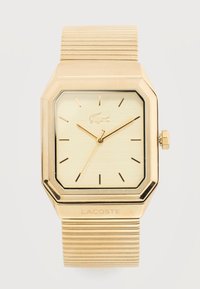 Ρολόι χειρός Lacoste σε χρυσό τόνο με τετράγωνο καντράν, ελάχιστους δείκτες ώρας, τρία χρυσά δείκτες και υφασμένη μεταλλική ελαστική μπάντα.