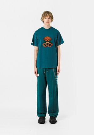 Camiseta de algodón en color verde aguamarina con un gráfico de un oso de peluche, combinada con pantalones verde aguamarina a juego que cuentan con un detalle de cadena lateral; ambos tienen un corte relajado.