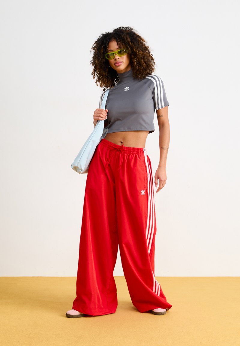 Top court gris avec des rayures blanches, pantalon large rouge avec des accents blancs, et un sac bleu clair. Le mannequin porte des lunettes de soleil vertes.
