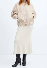 Beige fleece bomber jacket with a cropped fit and two front pockets, paired with a long cream ribbed skirt and black knee-high boots.