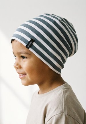 Young child smiling, wearing a gray and white striped beanie and a beige shirt, facing left against a plain white background.