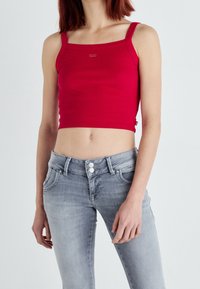 Femme portant un débardeur rouge court et un jean taille haute gris clair se tenant devant un fond uni.