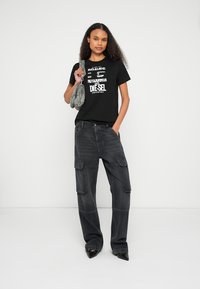 Μαύρο γραφικό t-shirt με λευκό κείμενο σε συνδυασμό με φαρδιά σκούρα γκρι παντελόνια cargo. Το μοντέλο κρατάει μια τσάντα με σχέδια, φορώντας μυτερά μαύρα παπούτσια.