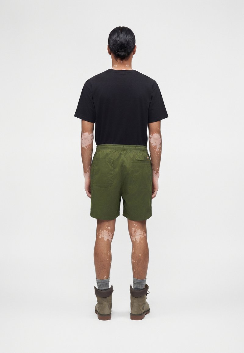 Person mit Vitiligo steht mit dem Rücken zur Kamera, trägt ein schwarzes T-Shirt, olivgrüne Shorts, graue Socken und braune Wanderschuhe vor weißem Hintergrund.